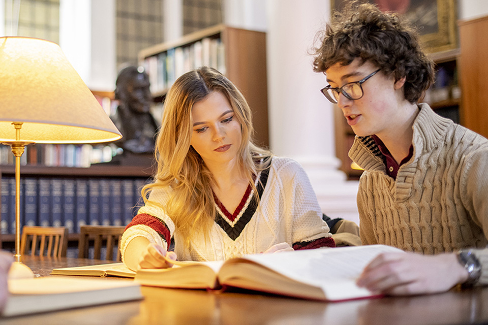 Two students reading books next to a small lit lamp, on a wooden table, surrounded by bookcases full of books and manuscripts in the background.test